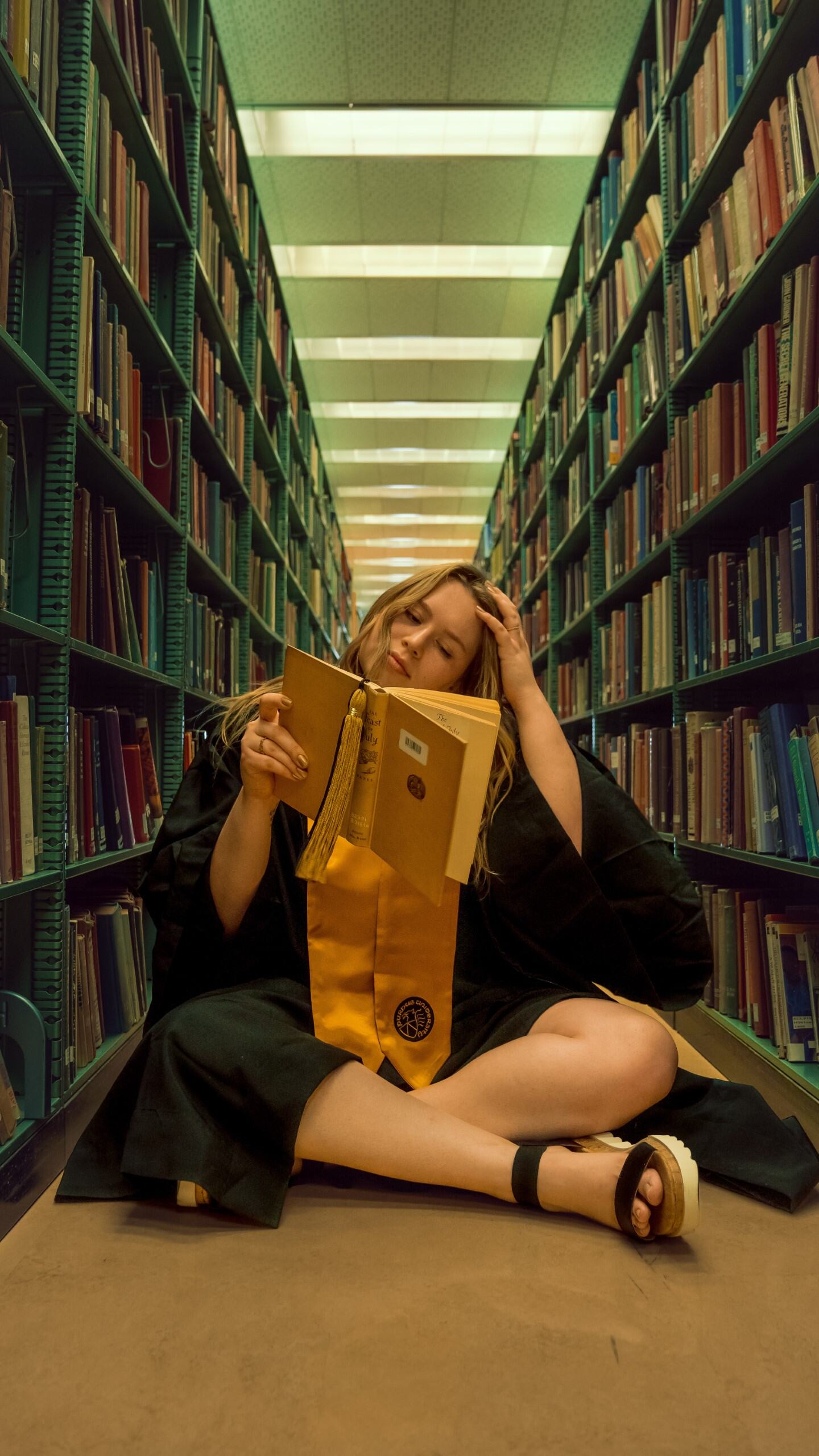 A student reading cross-legged on the floor between library shelves